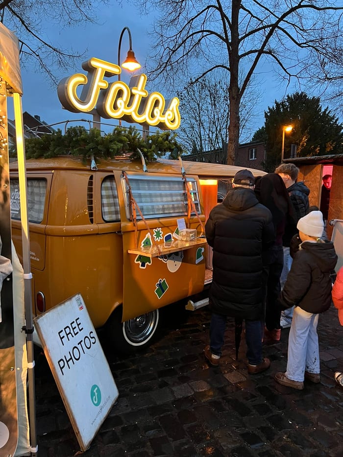 Abendliche Warteschlange vor dem gelben Foto Bulli auf einem Weihnachtsmarkt, darüber die leuchtende Fotos-Schrift.
