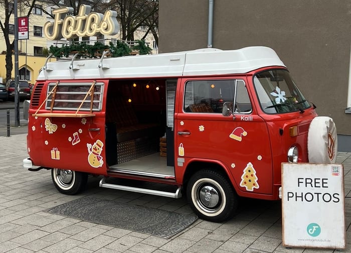 Roter VW-Bulli im Weihnachtsschmuck mit goldenen Aufklebern, Tannengirlande auf dem Dachträger und Leuchtschrift Fotos.