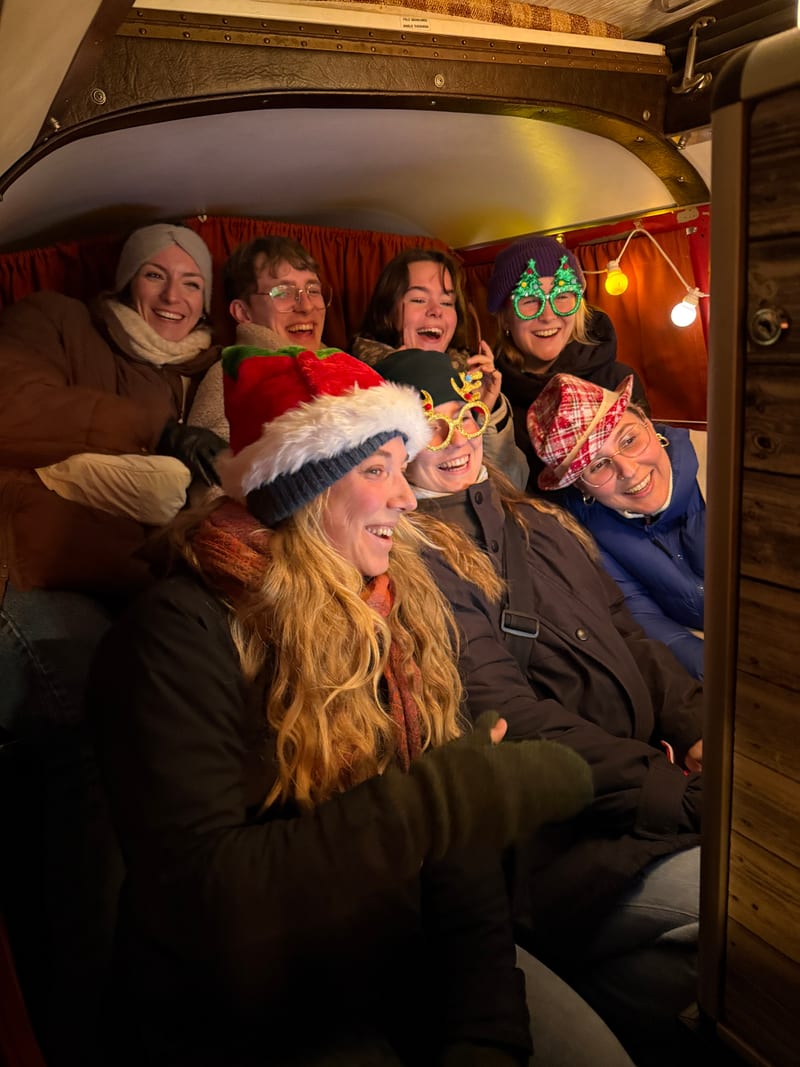 Sieben lachende Gäste mit Weihnachtsmützen und Spaßbrillen dicht gedrängt auf der Sitzbank im Foto Bulli.
