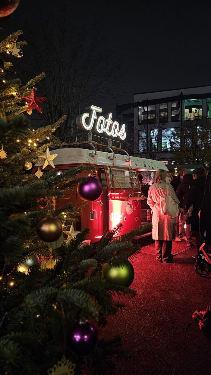 Blick durch einen geschmückten Tannenbaum auf den rot beleuchteten Foto Bulli bei Nacht, davor warten Marktbesucher.
