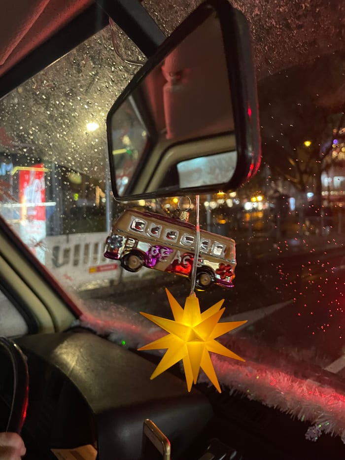 Blick aus dem fahrenden Tour-Bulli auf die abendliche Kölner Innenstadt, im Vordergrund ein leuchtender Weihnachtsstern und ein Bulli-Anhänger am Spiegel.
