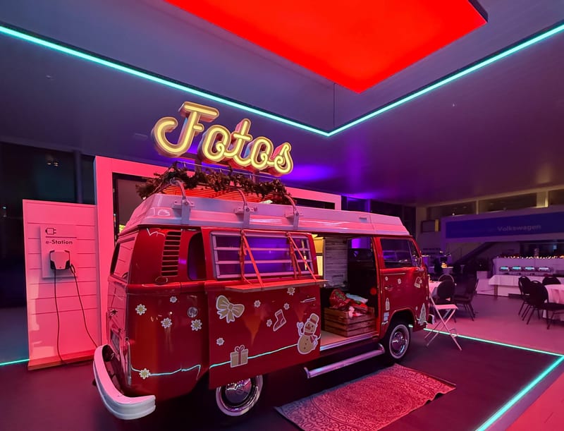 Roter Foto Bulli mit leuchtender Fotos-Schrift auf einer abendlichen Firmen-Weihnachtsfeier in einem Showroom, im Hintergrund eingedeckte Tische.