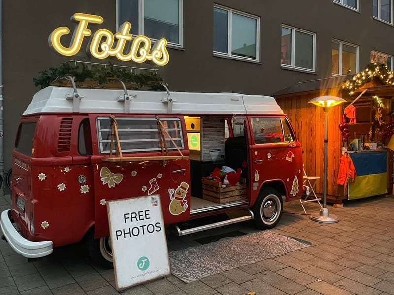 Roter Foto Bulli mit Tannengirlande und Leuchtschrift auf einem beleuchteten Weihnachtsmarktstand, daneben ein Aufsteller mit der Aufschrift Free Photos.